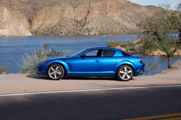 Blue Mazda RX-8 parked in profile beside a desert lake with red rock cliffs behind