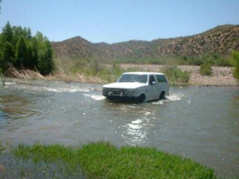 1993 Ford Bronco - 1993 Ford Bronco navigates a shallow river crossing under a clear blue sky.