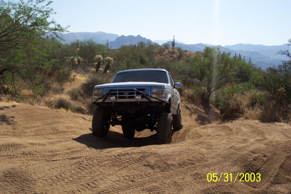 Lifted white Ford Bronco with black tube bumper on a sandy desert trail, cactus-covered hills behind