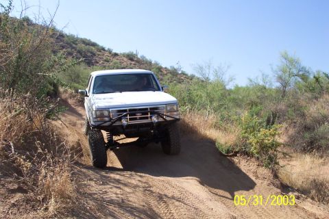 1993 Ford Bronco - A white Ford Bronco navigates a dusty, off-road trail under a clear blue sky.