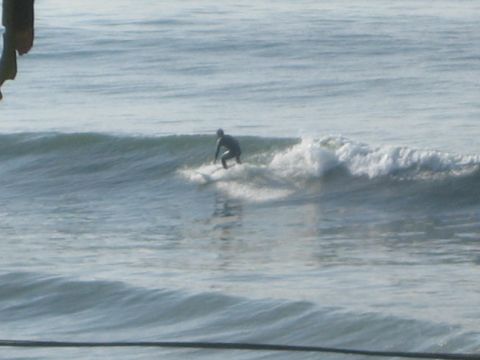 1990 Ford Bronco - wetsuited surfer rides along a small breaking wave, trailing whitewater across the gray-green ocean.