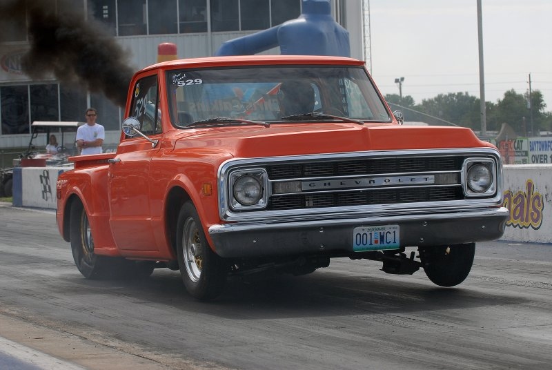 Orange classic Chevrolet stepside pickup lifting front wheels at a drag strip launch, race number 529 on windshield