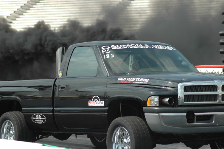 Black Dodge Ram diesel drag truck billowing black smoke at a drag strip, side/front view