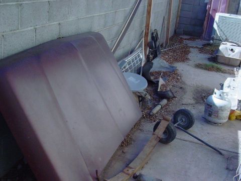 1979 Ford Bronco - A maroon car hood is stored outdoors next to a cinder block wall, surrounded by discarded vehicle parts and propane tanks.