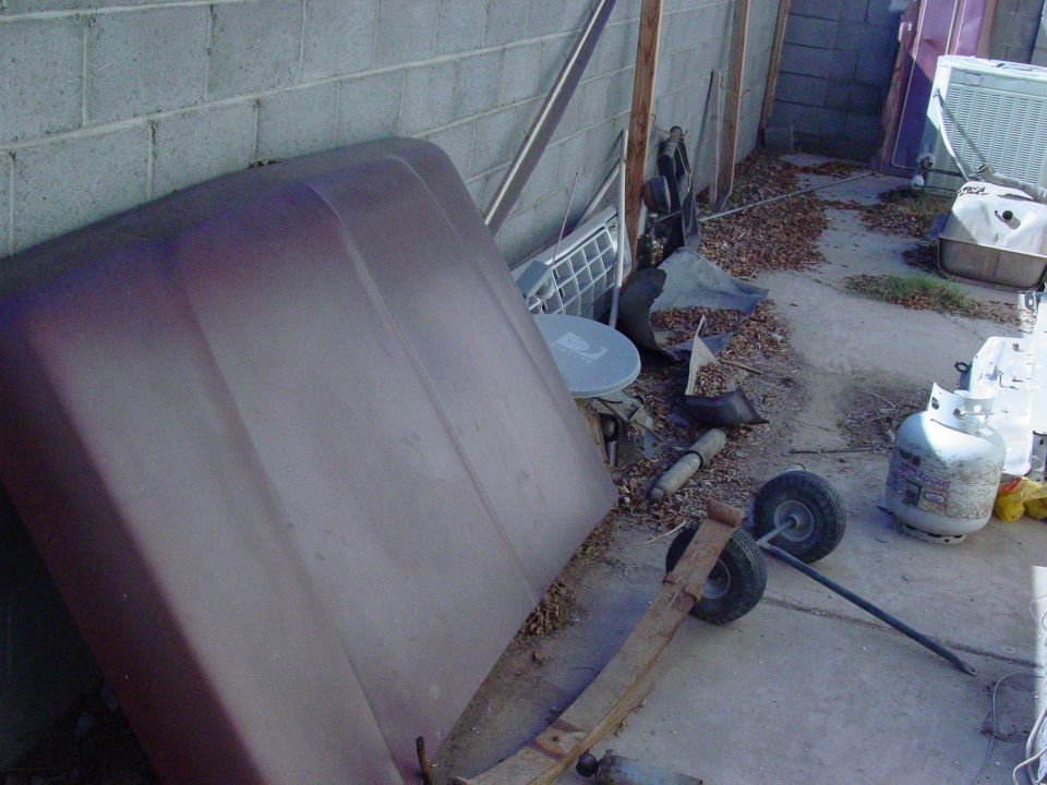 Maroon car hood leaning against a cinder block wall, with various parts and propane tanks scattered around