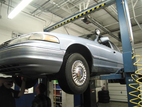 1991 Ford LTD Crown Victoria - A light blue sedan is raised on a car lift in a garage, with mechanics working underneath.