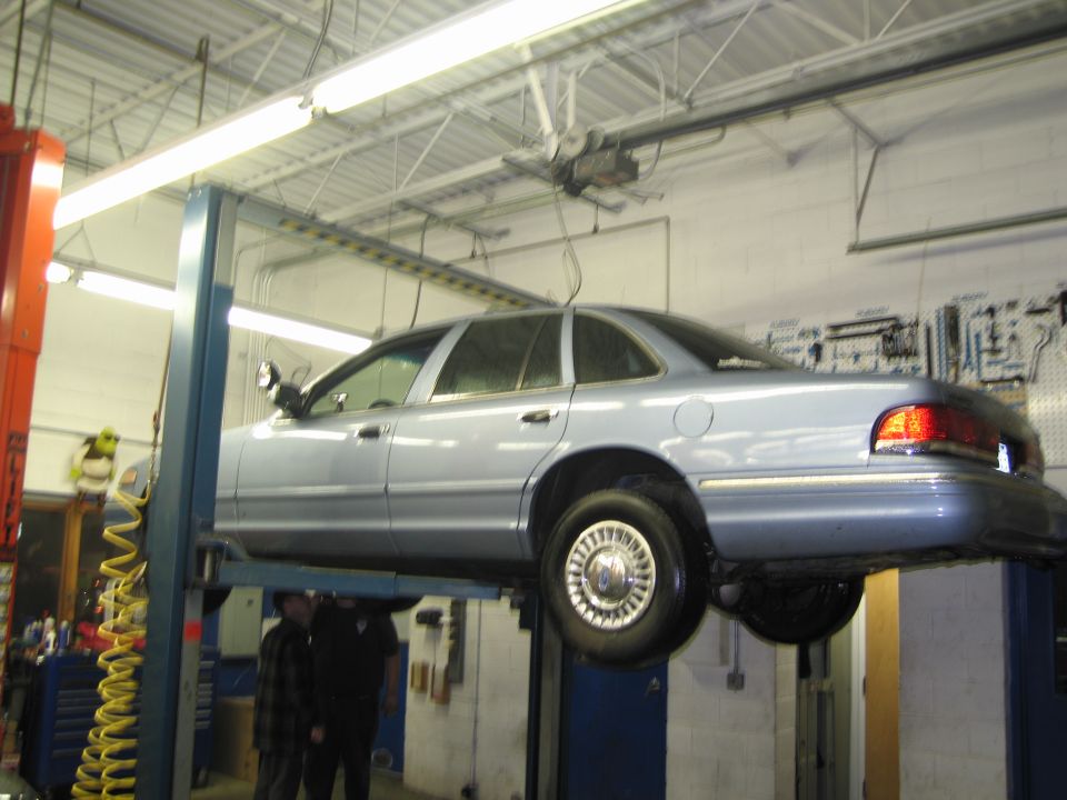 Light blue sedan on a car lift, viewed from the side. The car has a chrome trim and appears to be in a repair shop