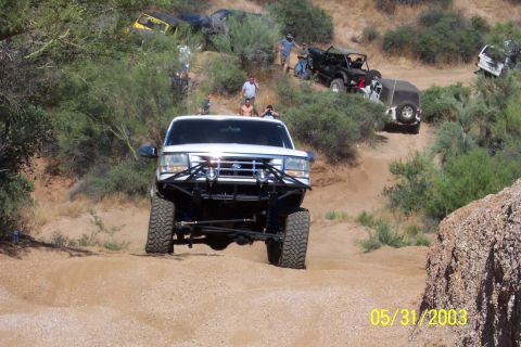 1993 Ford Bronco - A white Ford F-Series truck navigates a dusty off-road trail with other vehicles visible on the hillside.