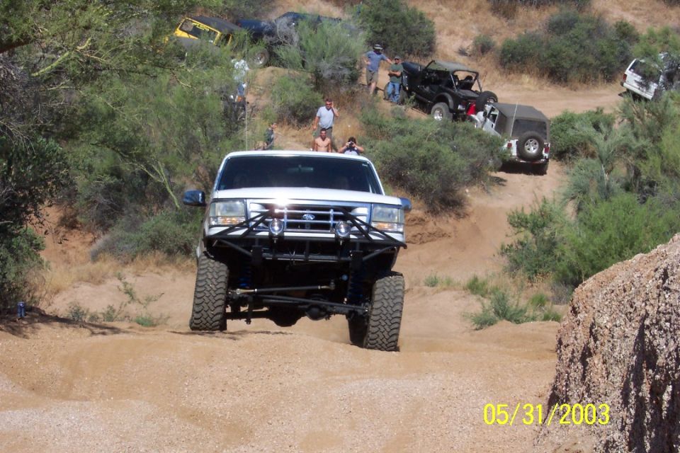 Lifted white Ford Bronco with brush guard climbing a sandy desert trail on big tires
