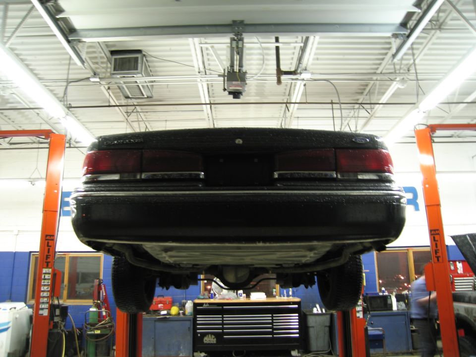 Rear view of a black sedan on a lift in a garage. The taillights and trunk are visible