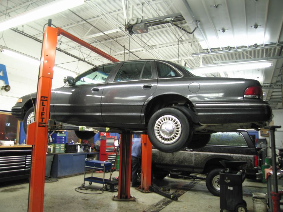 Dark gray sedan on a lift, side view. Another vehicle is below it. The car has a 1992 body style