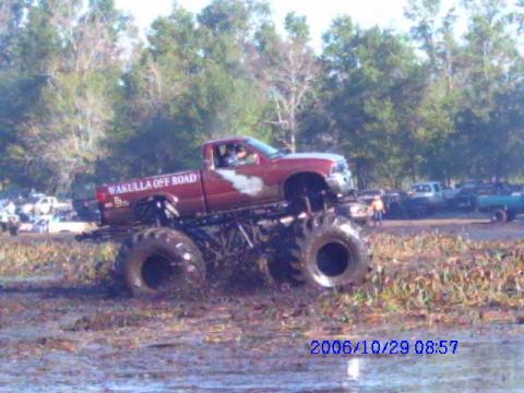 1987 Suzuki Samurai - A modified red truck with oversized tires navigates a muddy terrain, with other vehicles visible in the background.