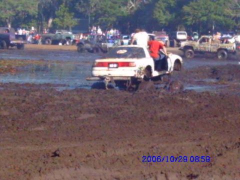 1987 Suzuki Samurai - A white SUV with a red stripe is stuck in the mud during an event, with other vehicles and spectators in the background.