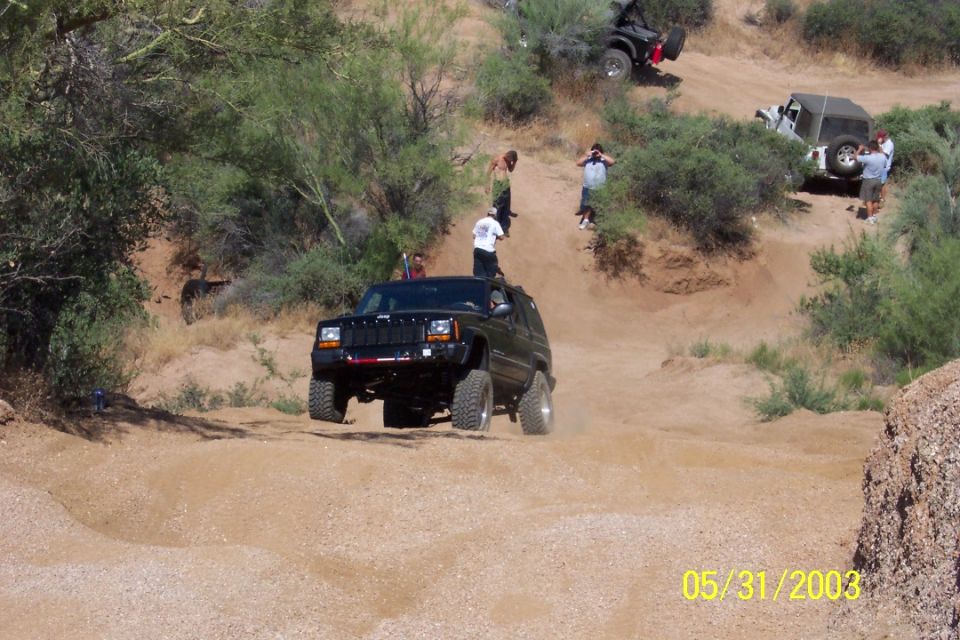 Dark green lifted Jeep Cherokee XJ on big tires in a sandy desert wash with onlookers