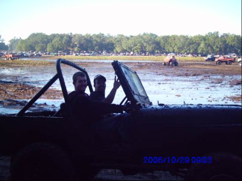 1987 Suzuki Samurai - Two people are in a muddy off-road vehicle, with other vehicles visible in the background at a mud bog event.