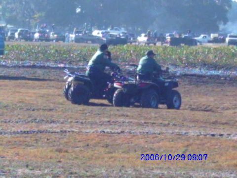 1987 Suzuki Samurai - Two riders on ATVs navigate a muddy terrain, with a crowd and vehicles visible in the distance.