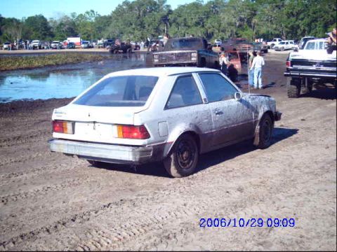 1987 Suzuki Samurai - A white coupe sits in the mud, with other vehicles and people in the background at an outdoor event.