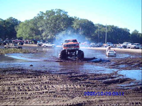 1987 Suzuki Samurai - A monster truck navigates a muddy track, spraying water as it goes, with spectators and other vehicles visible in the distance.