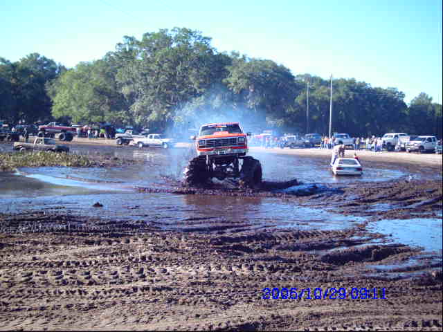 Monster truck driving through mud, kicking up water. Other vehicles and people in background