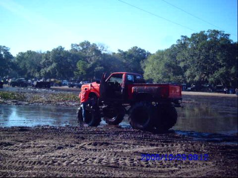 1987 Suzuki Samurai - A lifted red truck with large tires navigates a muddy terrain under a clear blue sky.
