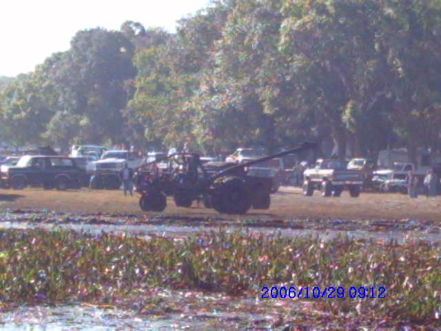 Off-road vehicle with crane arm in muddy field, multiple trucks in background