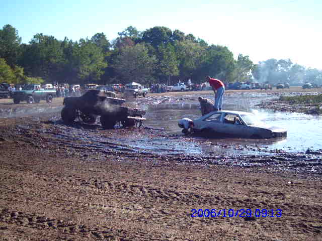 Mud bog event with several trucks and a car stuck in the mud. A person stands on the roof of the car