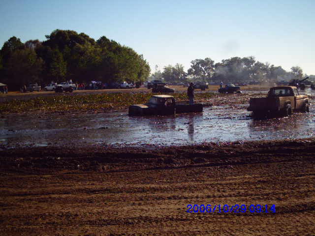 trucks stuck in a mud pit with trees and people in the background