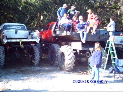 1987 Suzuki Samurai - Several people are gathered around two large monster trucks in an outdoor setting.