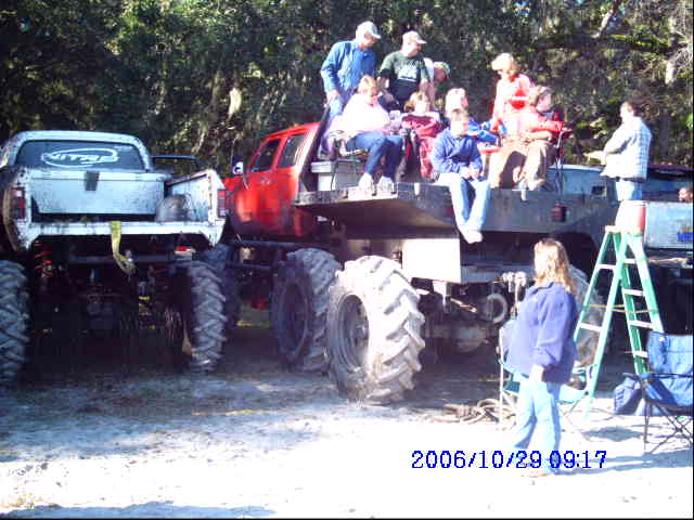 Two monster trucks parked side-by-side, one white and one red, with people riding on the red truck's flatbed