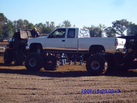 1987 Suzuki Samurai - A lifted pickup truck is parked next to another large vehicle on a dirt lot.