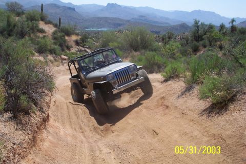 1993 Ford Bronco - A Jeep Wrangler navigates a steep, dusty trail surrounded by desert vegetation and mountains in the background.