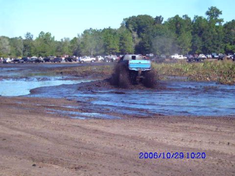1987 Suzuki Samurai - A blue pickup truck navigates a muddy track, kicking up water and mud as other vehicles wait on the bank.