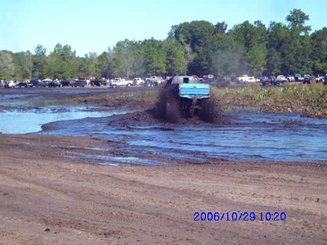 Blue pickup truck splashing through mud, with other vehicles parked in background