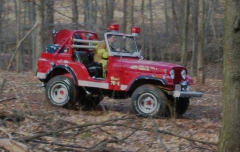 1989 Ford Bronco - A red Jeep CJ-series vehicle, modified as a fire truck, drives through a wooded area with a firefighter at the wheel.