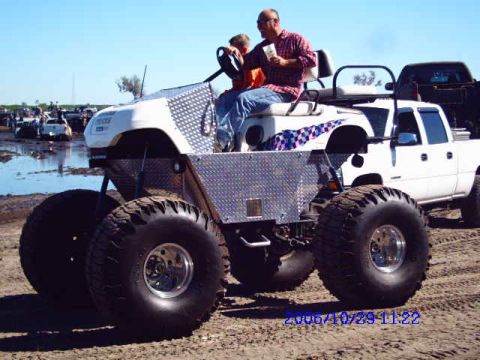 1987 Suzuki Samurai - A custom-built monster golf cart with oversized tires is driven by a man and a child, with a pickup truck visible in the background.