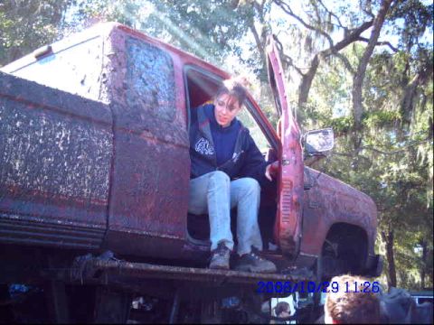 1987 Suzuki Samurai - Mud-covered red pickup truck with its door open, a woman seated inside, parked outdoors.