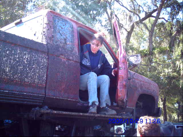 Red pickup truck covered in mud, driver's side door open, woman sitting inside