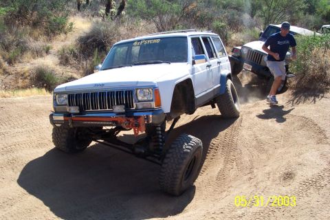 1993 Ford Bronco - A lifted white Jeep Cherokee XJ navigates a dusty off-road trail with another vehicle and a person in the background.