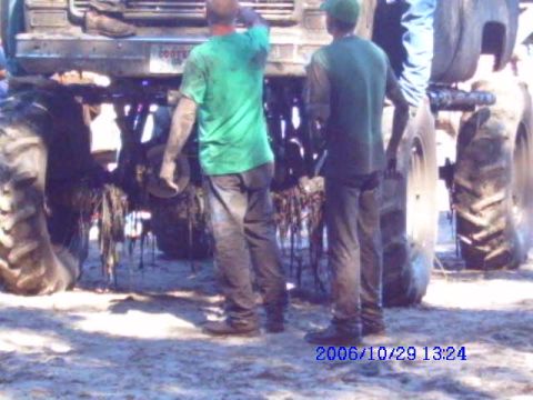 1987 Suzuki Samurai - Two men stand near the muddy undercarriage of a large monster truck with oversized tires.