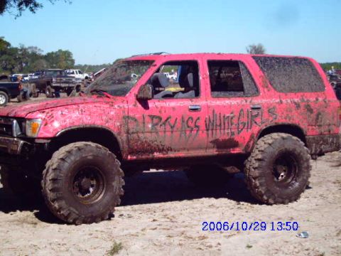 1987 Suzuki Samurai - A pink SUV, covered in mud, with large off-road tires and custom text on its doors, sits in a muddy field.