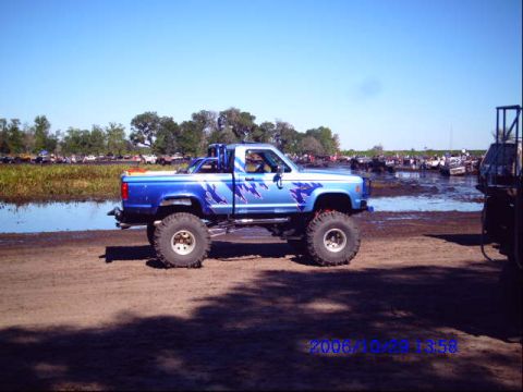 1987 Suzuki Samurai - A lifted blue pickup truck sits on a muddy bank next to a crowded waterway.