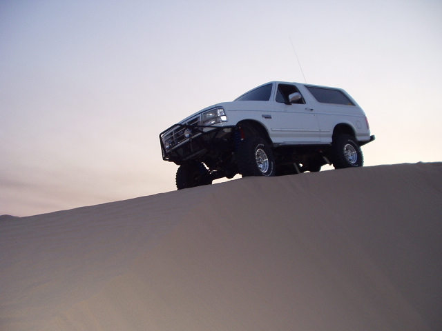 White lifted Ford Bronco SUV cresting a sand dune at dusk, low front-quarter view