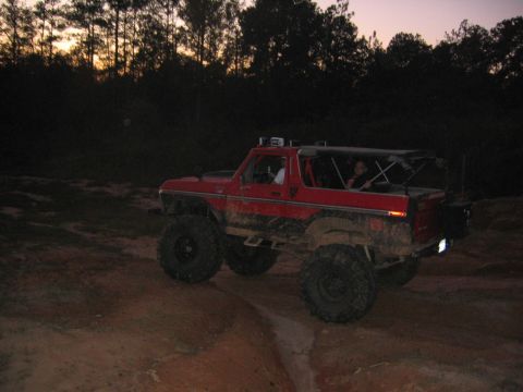1979 Ford Bronco - A red Ford Bronco with off-road modifications navigates a muddy trail at dusk.
