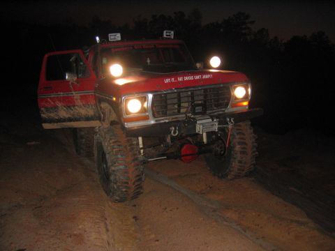 1979 Ford Bronco - A red 1979 Ford Bronco with its door open is shown off-road at night, equipped with auxiliary lights and a winch.