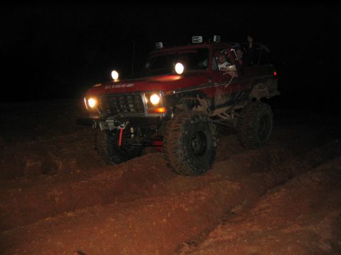 1979 Ford Bronco - A red off-road SUV navigates a dirt path at night, illuminated by its headlights and auxiliary lights.