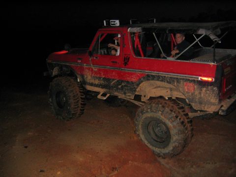 1979 Ford Bronco - Mud-covered red 1978 Ford Bronco with off-road tires and a soft top navigates a dirt path at night.