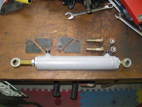 1981 Ford Bronco - A hydraulic steering cylinder and associated hardware are laid out on a wooden surface, possibly for installation or repair.