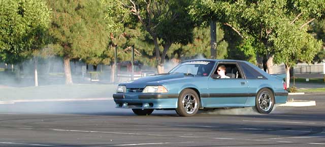 Light blue Fox-body Ford Mustang hatchback doing a burnout in a parking lot with tire smoke behind it