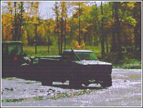 1976 Chevrolet Silverado 2500 - A pickup truck is stuck in the mud, with a tractor visible behind it. The scene is set against a backdrop of autumn trees.