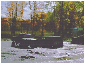 1976 Chevrolet Silverado 2500 - Several trucks are seen in a muddy area surrounded by trees with fall colors, under a SuperMotors logo.